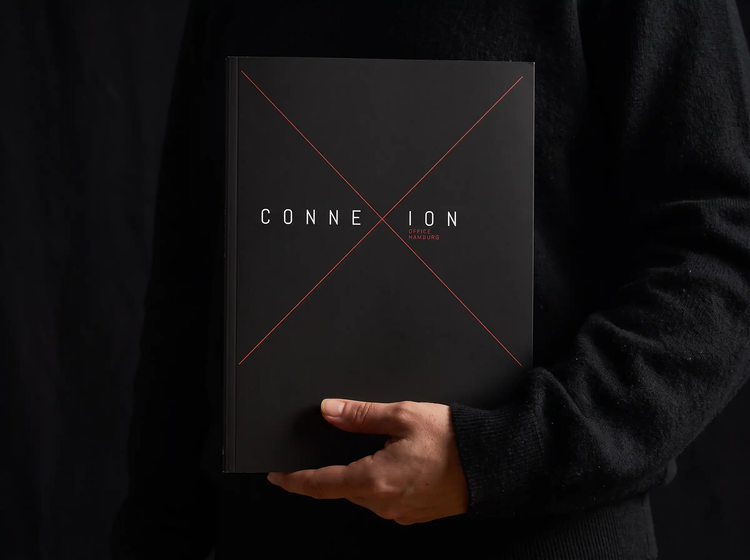 Schwarze ConneXion-Broschüre mit rotem X auf dem Cover, vor dunkler Kleidung gehalten