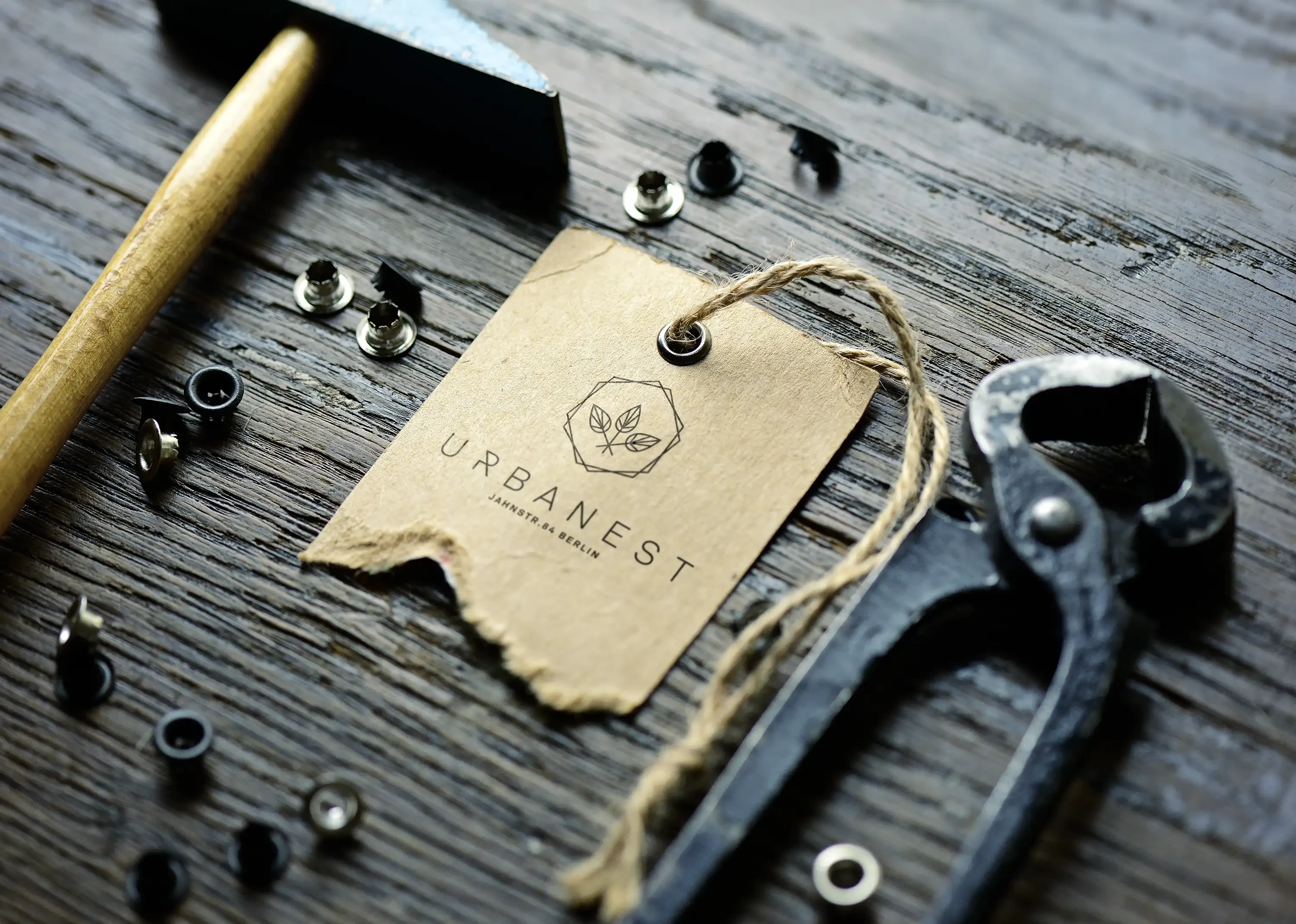 Urbanest-Anhänger aus Karton mit Logo zwischen Hammer, Zange und Ösen auf dunklem Holz